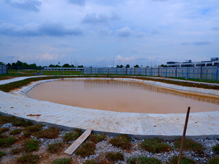 a horizontal wide-angle landscape shot capturing an oval stormwater retention pond lined with a concrete apron walkway, surrounded by construction hoarding and cloudy sky. © iwan33