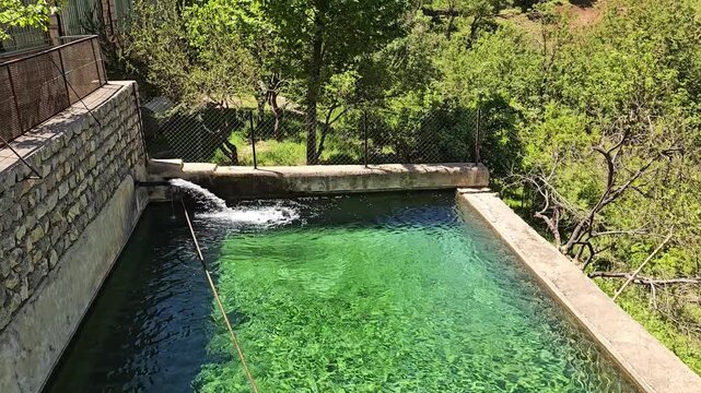 A vivid turquoise stone pool fed by flowing stream water rests below a rustic stone house, with a tall tree and rolling green hillsides under clear spring sky.
📍 AbbasAbad, Hawraman Takht, Kurdistan 
