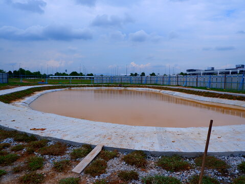 a horizontal wide-angle landscape shot capturing an oval stormwater retention pond lined with a concrete apron walkway, surrounded by construction hoarding and cloudy sky.