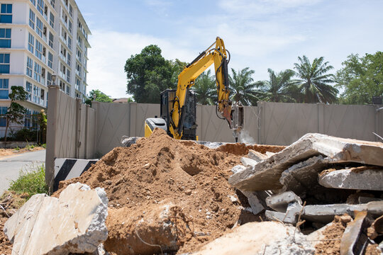 Excavator with hydraulic breaker working at demolition site near apartment complex and road