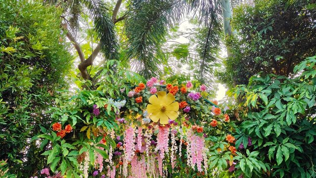 Colorful floral installation surrounded by lush tropical trees and hanging blossoms, creating a vibrant outdoor garden setting for weddings, events, and celebrations.