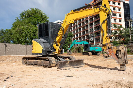 Yellow excavator with hydraulic breaker attachment and bulldozer blade parked at urban construction site