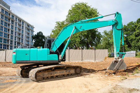 Green Tracked Excavator Digger Machine Parked On Sandy Ground At Urban Construction Site In Front Of Building