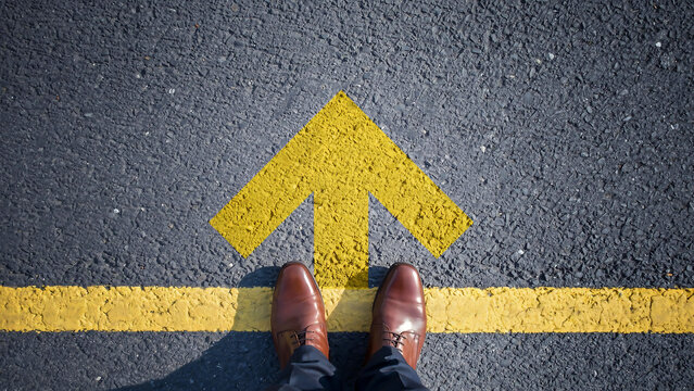 Person standing on asphalt looking at a painted yellow arrow pointing forward with direction decision business motivation standing guidance leadership planning strategy movement