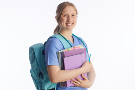 A young nursing student in blue scrubs and a stethoscope smiles while carrying a turquoise backpack and a stack of notebooks, ready for her clinical training session.
