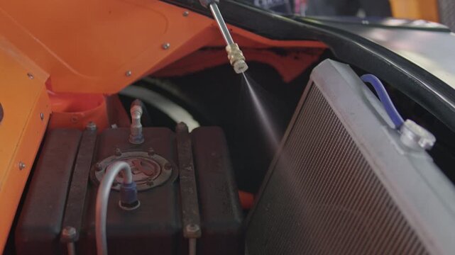 Close-up action shot of a spray nozzle washing down the front radiator and custom black fuel cell inside the engine bay of a race car.