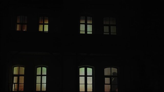Silhouette of a man with a cigarette in the window of an apartment building. A persons silhouette stands in a warmly lit window of a dark house at dusk