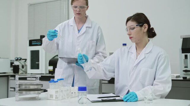 Focused female scientists teamwork examining chemical solution in flask and writing research report on clipboard in sterile cleanroom laboratory for medical pharmaceutical biotechnology innovation.