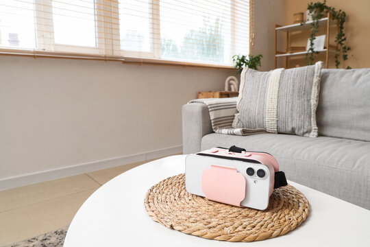 Pink and white virtual reality headset with a smartphone on a woven mat in a modern living room, the modern living room features a gray sofa with a textured pillow and blanket, a window with blinds