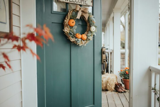 A welcoming teal front door adorned with an autumn-themed wreath featuring small orange and white pumpkins a natural burlap bow and dried leafy green accents for fall decoration