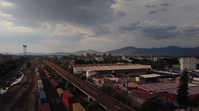 4K aerial drone footage flying forward over an industrial area with freight railway tracks, warehouses and factories under a cloudy afternoon sky.
