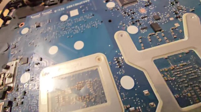 A close-up view of a blue laptop motherboard showcasing electronic components, microchips, printed circuits, and heat sink mounts during internal hardware inspection and repair.
