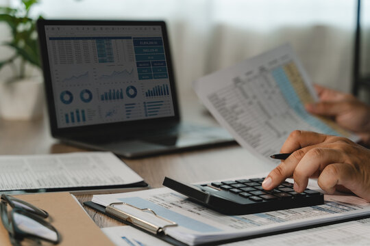 Investment Consultant reports with a calculator while a laptop displays charts and metrics, showing data review and business planning workflow for accounting and report evaluation.