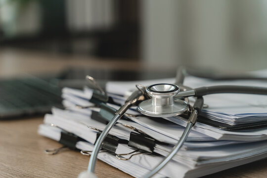 Stethoscope rests on clipped stacks of medical paperwork across on desk, document organization, records filing, healthcare administration workflow copy space.