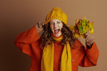 Obraz premium A woman in a yellow knit hat, orange sweater, and scarf playfully shouts with eyes closed, fingers in ears, holding autumn leaves. Against a brown background, she joyfully reacts to the fall season.