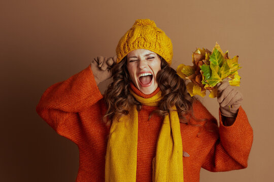 A woman in a yellow knit hat, orange sweater, and scarf playfully shouts with eyes closed, fingers in ears, holding autumn leaves. Against a brown background, she joyfully reacts to the fall season.