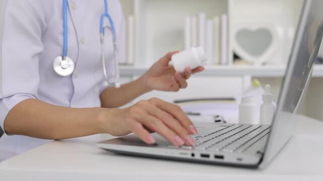Female doctor typing on laptop while holding medicine bottle.