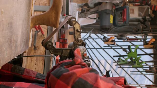 Vertical video. Carpenter equipment. Sanding wood. Man polishing figure piece product with handheld belt sander in joinery with professional tool kit on wall.