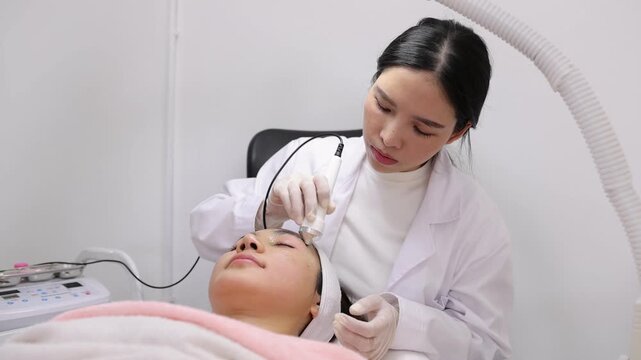 Young Asian beautician using a cosmetic device for a rejuvenating facial treatment on a client relaxing in a modern beauty spa salon.