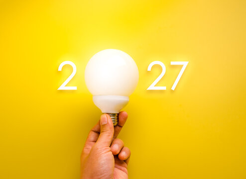 Human hand holds a glowing white globe light bulb forming the zero in 2027 against a vivid yellow background, symbolizing fresh ideas, innovation, inspiration, planning and a bright future concept.