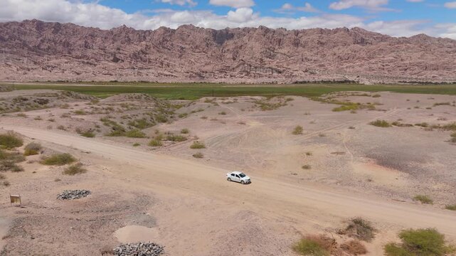 Drone tracking white car on desert road approaching Quebrada de las Flechas mountains, aerial medium pullback at angle