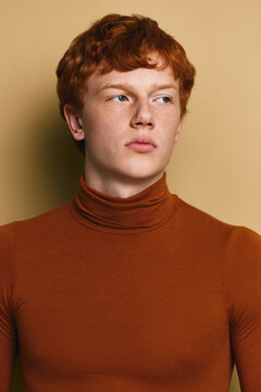 Young man with red hair and freckles wearing brown turtleneck sweater looks sideways with serious expression on beige background. Portrait of confident male model.