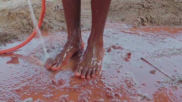 Close up of a native person cleaning their bare feet covered in clay. The water from a hose rinses away the mud after working