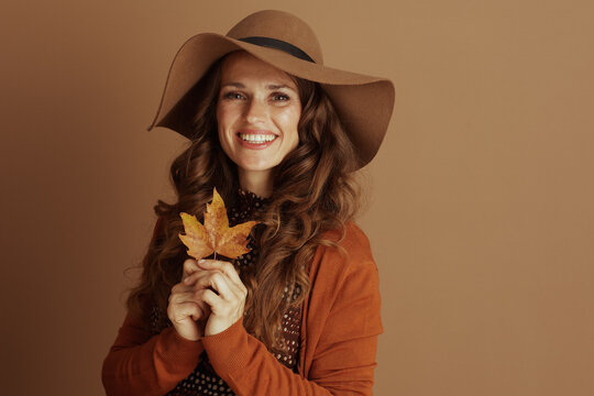 A smiling woman with curly brown hair, dressed in autumnal fashion with a wide-brimmed hat and orange cardigan, holds a yellow leaf against a warm brown background.