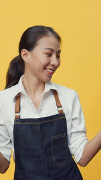 Vertical Screen : Happy smiling Asia waitress wearing apron friendly looking at camera giving coffee or tea and credit card reader machine to customer isolated on yellow background. Entrepreneur cafe.