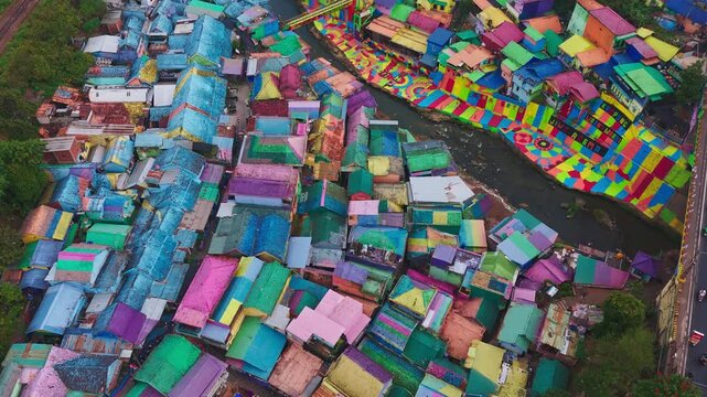 Colorful rooftops dominate this vibrant slum settlement. Winding paths cut through densely packed structures. Muddy river runs alongside the crowded dwellings. Buildings show varied hues