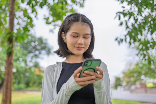 Asian Gen Z woman using AI fitness tracking app on smartphone before jogging in green urban park.