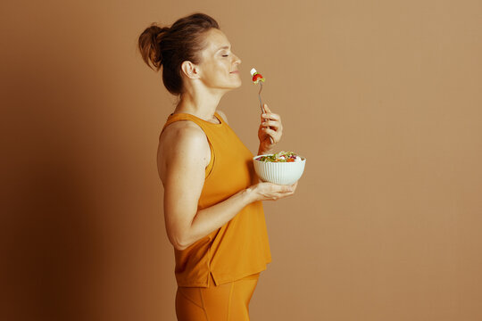 In a side profile, a woman with closed eyes, dressed in an orange athletic outfit, smells a forkful of fresh salad. This image conveys the essence of mindful eating and appreciation for healthy food.