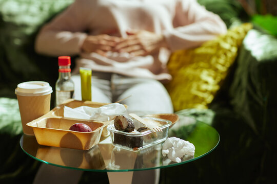 Sitting on a green couch, a woman clutches her midsection in pain after consuming a takeaway meal. Leftover sushi and containers sit on the table, reflecting poor dietary choices.
