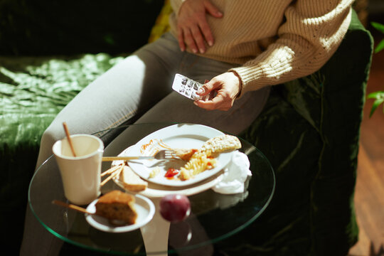 Resting on a green sofa, a woman holds her stomach in pain after a meal of pasta and cake, preparing to take medication for digestive relief.