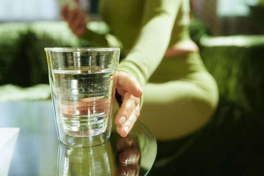 A close-up of a woman in sage green activewear resting her hand near a glass of fresh water on a glass table, emphasizing daily hydration and a balanced wellness routine at home.