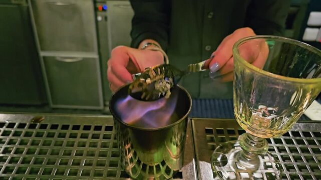 Bartender pours fire from a metal mixing cup into an empty goblet during cocktail preparation, with blue flames, orange glow, steel drip tray, and moody bar lighting at night.
