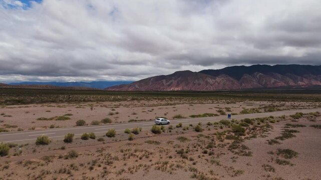 Drone tracking car along Recta del Tin Tin beneath dramatic mountain clouds, aerial angled follow orbit