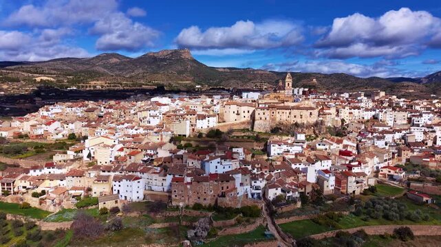 Panoramic view of Chelva, a traditional Spanish village in Valencia region, featuring white and terracotta rooftops, a historic church tower, and mountain backdrop under blue skies