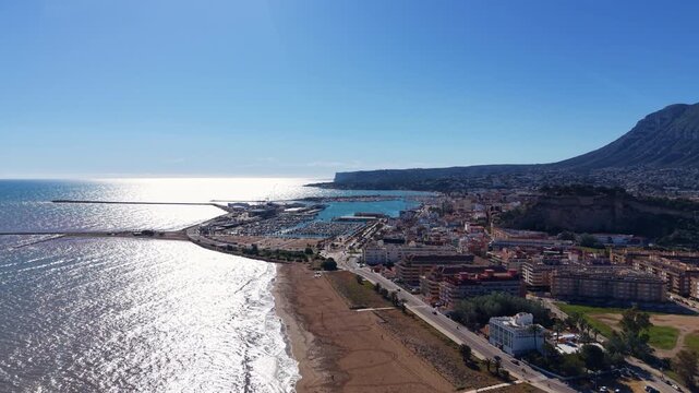 Panoramic view of Denia, Spain, revealing a sandy beach, marina with boats, coastal town buildings, and a dramatic rocky mountain under a clear blue sky