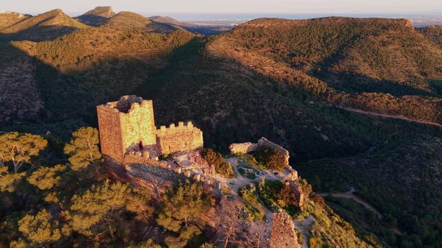 Medieval castle ruins of Serra atop a rocky hilltop in Sierra Calderona natural park, Spain, bathed in warm golden sunset light surrounded by forested mountains