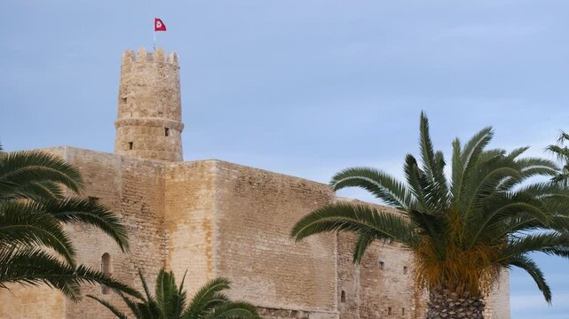 Ribat of Monastir in Tunisia with stone fortress walls and watchtower flying national flag, framed by palm trees under dramatic sky, highlighting historic Islamic architecture