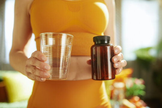Close-up of a woman in orange sportswear holding a glass of water and a bottle of supplements, emphasizing hydration and daily wellness habits.