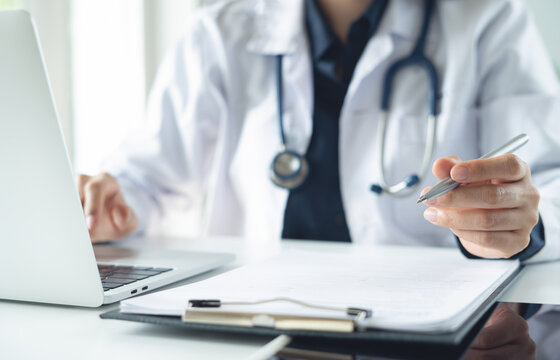 Close up, doctor working on laptop at medical clinic and writing medical reports, searching patient information on electronic health record with medical document on table
