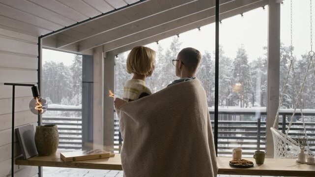 Caucasian adult man and woman wrapped in blanket standing by window of winter cabin, hugging and looking at snowy forest. Ideal for winter travel, real estate, or rental promotion