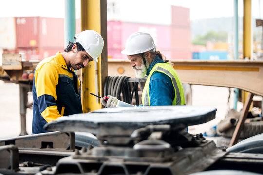 Two industrial workers in hard hats inspect a truck"s fifth-wheel coupling in a workshop at a shipping depot, using tools and safety gear amid containers and heavy equipment.