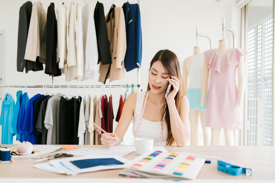 Fashion designer in a bright studio on the phone, sketching with color swatches and fabric samples; garments hang on racks and mannequins display dresses.