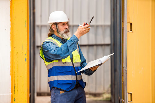 Safety-vested site worker using a walkie-talkie and checking paperwork between cargo containers at an industrial yard.