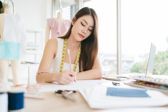 Fashion designer in a bright studio sketches clothing designs at a desk, measuring tape around neck, mannequin and fabrics in the background.