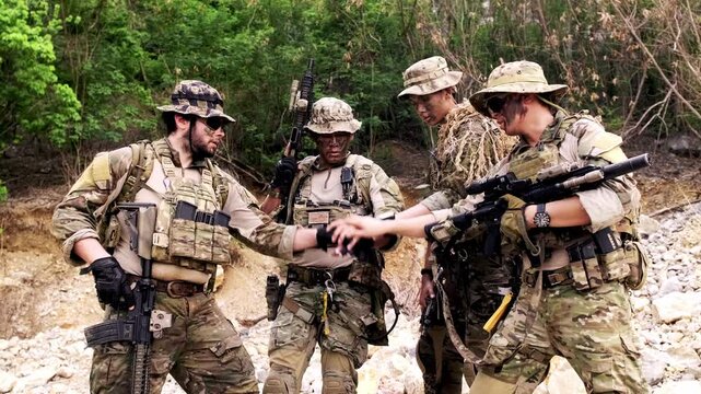 Four camouflaged soldiers in tactical gear form a team huddle outdoors, holding rifles during field training in a rugged, wooded area.