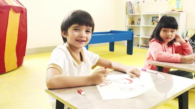 Smiling child draws with markers at a classroom desk while another student concentrates nearby; bright playroom with bookshelves and a game table